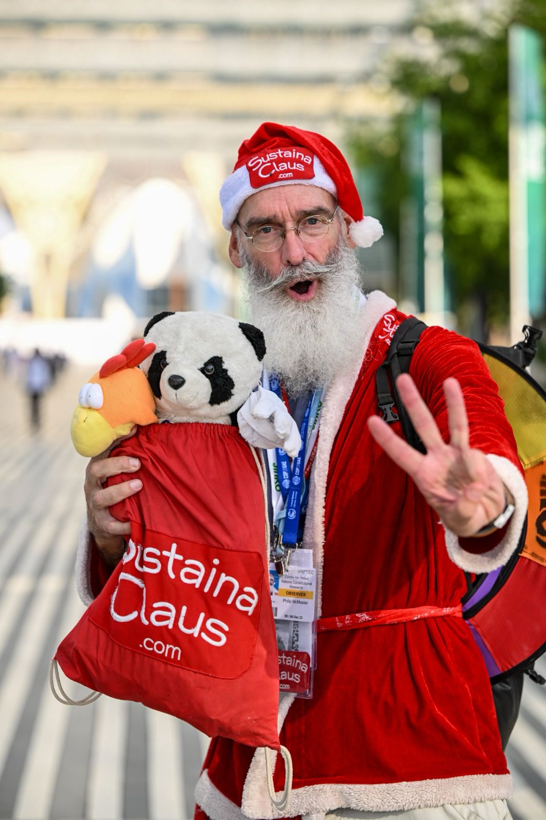 Philip McMaster as SustainaClaus at COP28, holding a SustainaClaus bag and a panda, raising three fingers